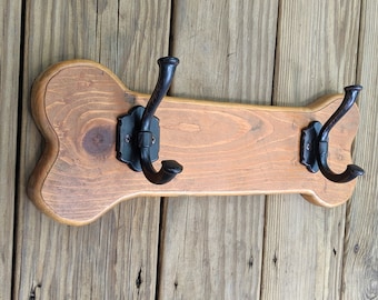 Può includere: Un gancio da parete in legno a forma di osso di cane con due ganci in metallo nero. Il legno ha una finitura naturale con venature visibili. I ganci sono montati su una placca decorativa.