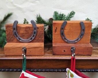 Può includere: Due fermalibri rustici in legno con un design a ferro di cavallo. Ogni fermalibri presenta un ferro di cavallo in metallo grigio attaccato a un blocco di legno rettangolare. Il legno ha un caldo tono marrone-rossastro. Il verde è visibile sullo sfondo.