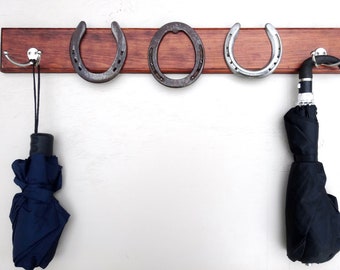 Può includere: Un attaccapanni da parete in legno con tre decorazioni a ferro di cavallo in metallo e due ganci doppi. Due ombrelli neri arrotolati sono appesi ai ganci. Il legno è tinto di marrone scuro.