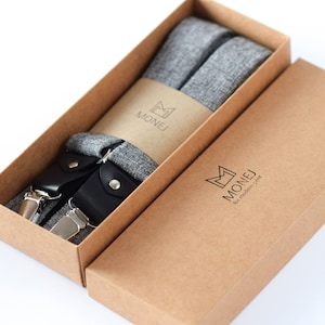 May include: A pair of gray suspenders with black leather accents and silver metal clips, packaged in a brown cardboard box with the brand name "MONEJ for modern gent" printed on the box.