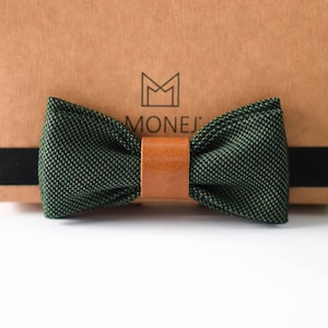 Può includere: Un papillon verde con un cinturino in pelle marrone. Il papillon è su una scatola marrone con la parola "MONEJI" stampata su di essa.