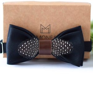 May include: A black bow tie with a brown leather strap and a pheasant feather design. The bow tie is on a brown box with the text "MONEF" on it.