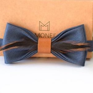 May include: A navy blue bow tie with brown feathers and a brown leather strap. The bow tie is on a brown box with the word "MONEJI" printed on it.