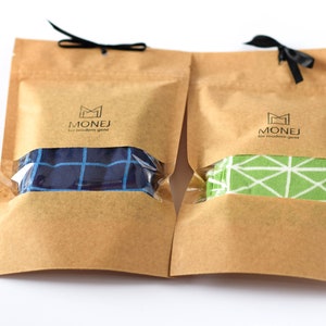 May include: Two brown paper bags with black ribbon ties. Each bag contains a pair of socks. One pair is blue with a white grid pattern, and the other pair is green with a white geometric pattern. The bags have a logo that says "MONEJ for modern gent".
