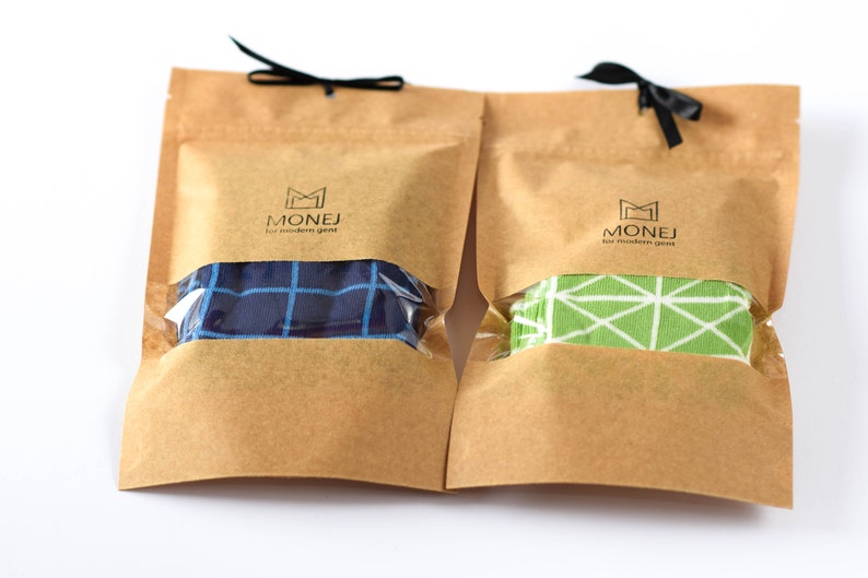 May include: Two brown paper bags with black ribbon ties. Each bag contains a pair of socks. One pair is blue with a white grid pattern, and the other pair is green with a white geometric pattern. The bags have a logo that says "MONEJ for modern gent".