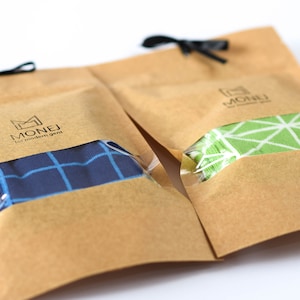 May include: Two brown paper bags with black ribbon ties. Each bag contains a pair of socks. One pair is blue and white checkered, the other is green and white geometric. The bags have the logo "MONEJ for modern gent" printed on them.
