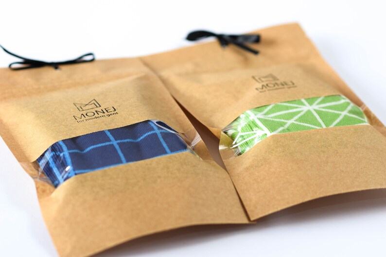 May include: Two brown paper bags with black ribbon ties. Each bag contains a pair of socks. One pair is blue and white checkered, the other is green and white geometric. The bags have the logo "MONEJ for modern gent" printed on them.