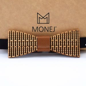 Peut inclure: Un nœud papillon en bois avec un motif de grille découpé au laser et une bande centrale en cuir marron lisse. Le nœud papillon est fixé à une sangle élastique noire et est placé devant une boîte en carton beige avec le logo MONEJ imprimé en noir.