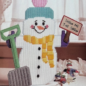May include: A plastic canvas snowman decoration featuring a light blue hat with a pink pom-pom, a yellow scarf, and three black buttons. The snowman holds a green shovel and a sign that reads SHOW ME THE SNOW in black letters on a red background.