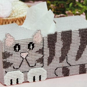 May include: Gray and white crocheted tissue box cover with a cat design. The cat has pink ears, a gray nose, and black eyes. The cover is shaped like a cat lying down.