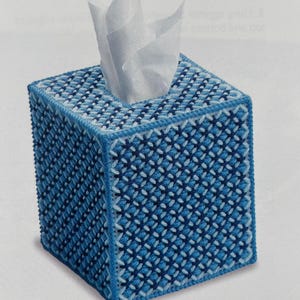 May include: A square tissue box cover in shades of blue, with a cross-stitch pattern. The cover has a light blue border and a square opening at the top for tissue dispensing. White tissues are visible.
