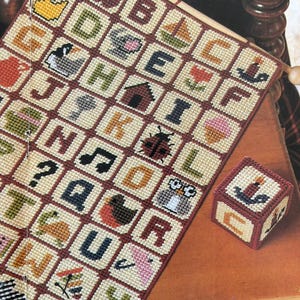 K&ouml;nnte beinhalten: Ein Kreuzstich-Alphabet-Sampler mit farbenfrohen Bildern und Buchstaben, darunter eine Ente, ein Apfel und ein Segelboot. Daneben liegt ein kleiner, passender W&uuml;rfel mit einem Kerzenbild. Der Sampler wird auf einem Holzstab pr&auml;sentiert.