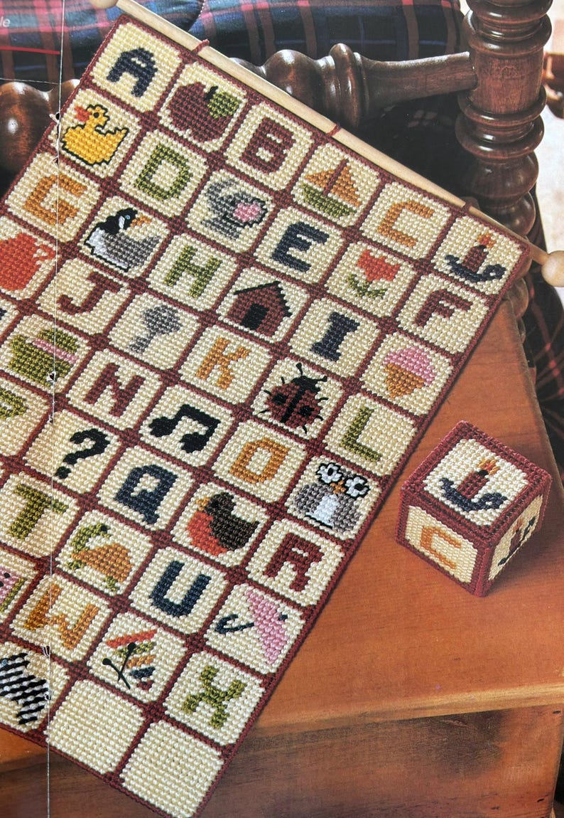 K&ouml;nnte beinhalten: Ein Kreuzstich-Alphabet-Sampler mit farbenfrohen Bildern und Buchstaben, darunter eine Ente, ein Apfel und ein Segelboot. Daneben liegt ein kleiner, passender W&uuml;rfel mit einem Kerzenbild. Der Sampler wird auf einem Holzstab pr&auml;sentiert.