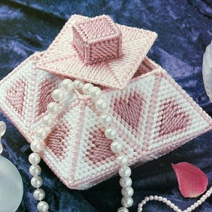 May include: A handmade pink and white needlepoint jewelry box with a matching lid and small square top. A pearl necklace is draped over the box. A perfume bottle and a rose petal are in the background.