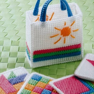 Puede incluir: Una bolsa de lona blanca con asas azules y un diseño de sol. La bolsa está rodeada de cuatro posavasos cuadrados coloridos con patrones geométricos.
