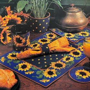 May include: A set of blue and yellow sunflower-themed placemats, coasters, and a mug. The placemats and coasters feature a repeating pattern of sunflowers with green leaves on a blue background. The mug has a similar design, with a sunflower pattern on a white background.