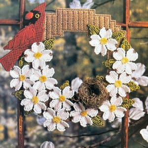 May include: A red cardinal bird perched on a wreath made of white flowers and green leaves. The wreath is decorated with a brown woven nest.
