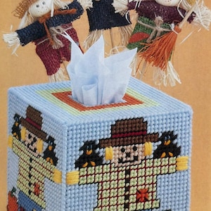 May include: A blue and white tissue box cover with a cross-stitch design of a scarecrow standing on pumpkins. Three small scarecrows are on top of the box.