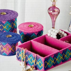 May include: Three pink and purple plastic canvas boxes with lids and a larger pink plastic canvas box with dividers. The boxes have a geometric pattern in shades of blue, purple, pink, and yellow.