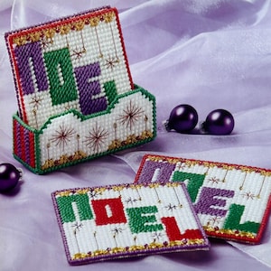May include: Set of four white canvas coasters with the word "NOEL" in green, purple, and red letters. The coasters have a gold glitter border and are decorated with white stars. The coasters are in a white plastic holder with a red, white, and purple striped design.