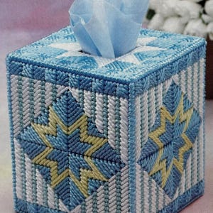 May include: A blue, white, and yellow tissue box cover with a starburst pattern. The cube-shaped cover is made from a woven material. A blue tissue is visible at the top. The design is handmade and decorative, with measurements in centimetres.