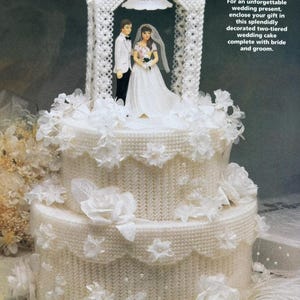 May include: A two-tiered white wedding cake with a white crocheted archway on top. The cake is decorated with white flowers and pearls. A bride and groom figurine are standing under the archway.