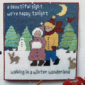 May include: A square plastic canvas needlepoint featuring a winter scene with two figures, a snowman, trees, a rabbit, and a bird. The text reads: a beautiful sight we're happy tonight walking in a winter wonderland. The border is dark red.