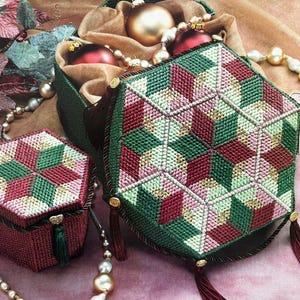 Könnte beinhalten: Zwei sechseckige Schachteln aus Stickstoff mit einem geometrischen Würfelmuster in Dunkelrot, Waldgrün und Blassrosa. Die Deckel sind mit goldfarbenen Akzenten und dunkelgrünen Quasten an den Ecken verziert.