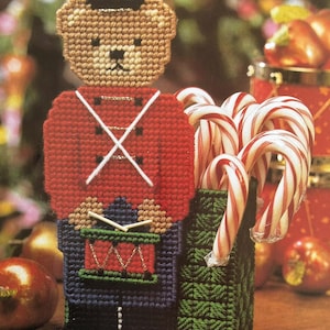 May include: A handmade Christmas decoration featuring a teddy bear soldier holding a drum and a green container filled with candy canes. The bear wears a red jacket, black hat, and blue pants. The candy canes are red and white.