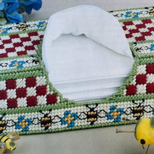 May include: A crocheted tissue box cover with a checkered pattern in red and white, green and white borders, and a bee motif. The cover is decorated with blue flowers and yellow bees.
