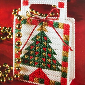 Puede incluir: Una bolsa de regalo de Navidad de lona blanca con un diseño de árbol de Navidad bordado en rojo, verde y dorado. La bolsa tiene una cinta roja atada alrededor del asa.