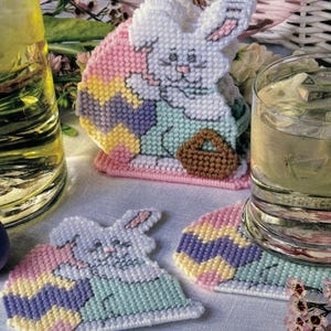 May include: Three crocheted Easter bunny coasters. The coasters are shaped like bunnies holding Easter eggs. The eggs are decorated with pastel colors and stripes. The bunnies are white with pink ears and brown eyes.