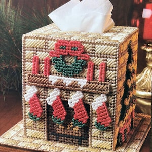 May include: A square plastic canvas tissue box cover designed to look like a brick fireplace. The front features a mantel with four red stockings, a green wreath with a red bow, and four red candles. A white tissue is pulled through the top opening.