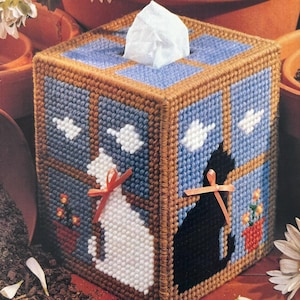 May include: A brown, blue, and white plastic canvas tissue box cover with a window scene featuring two cats, one white and one black, with red bows. The cats are looking out the window at two white birds.