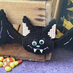 May include: A black felt bat with purple stitching and pink ears. The bat has googly eyes and a toothy grin. It is a Halloween decoration.
