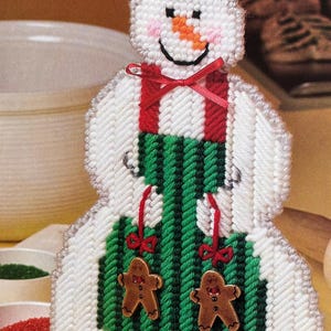 May include: A plastic canvas snowman figure wearing a white chef hat with a black band, a green and black striped apron with red straps, and two small brown gingerbread man buttons attached to the front with thin red ribbons.