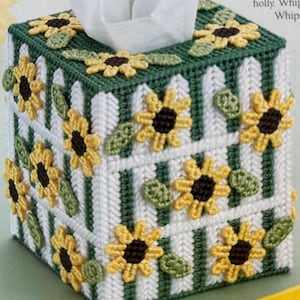 May include: A green and white tissue box cover with a white picket fence design and yellow sunflowers. The cover is made of plastic canvas and is decorated with yarn.