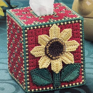 May include: A handmade tissue box cover featuring a vibrant sunflower design. The box is primarily red with a green and cream border. The sunflower has yellow petals and a dark brown center. White tissues are visible.