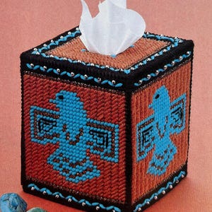 May include: A square tissue box cover with a Native American design featuring two blue birds with geometric patterns on a brown background. The cover is decorated with silver beads around the edges.