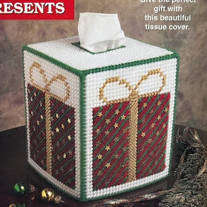 May include: A square plastic canvas tissue box cover designed to look like a wrapped gift. The cover features white edges, red and green diagonal striped panels, gold ribbon accents, and gold star embellishments. Text reads: RESENTS and Give the perfect gift with this beautiful tissue cover.
