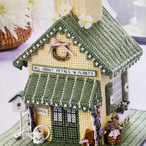 May include: A yellow and green tissue box cover shaped like a house with a sign that reads "All About Petals & Plants". The house has a green roof, white trim, and a green awning. There is a small birdhouse, a watering can, and a teddy bear in front of the house.