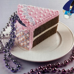Pode incluir: Uma fatia de bolo de crochê com cobertura rosa e detalhes decorativos brancos e azul claro está em um prato branco. O bolo tem uma base marrom com uma faixa branca. Colares de contas roxas e prateadas estão drapeados ao redor do prato.