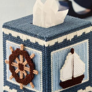 May include: A blue and white tissue box cover with a nautical theme. The cover features a brown ship's wheel, a white sailboat, and a blue and white striped background.