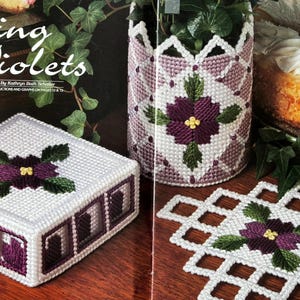 Può includere: Un set di artigianato in tela plastica intitolato Spring Violets di Kathryn Beth Schaller, composto da una scatola quadrata con coperchio, un contenitore cilindrico e un sottobicchiere quadrato, decorati con motivi di violette viola e foglie verdi.