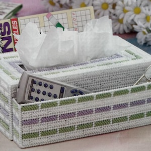 May include: A rectangular tissue box with a matching storage compartment, crafted with a white, green, and purple woven pattern. The box holds tissues, a remote control, reading glasses, and a crossword puzzle book.