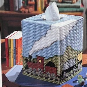 Puede incluir: Una funda para caja de pañuelos azul y blanca con un diseño de tren y nube. El tren es rojo, negro y gris. La nube es blanca. La funda para caja de pañuelos está hecha de lona plástica.