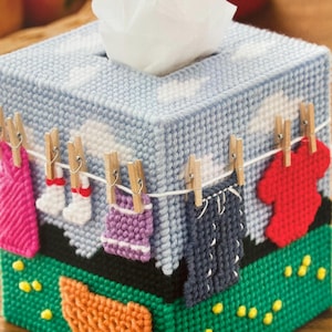 Op de afbeelding: Een blauwe en witte tissueboxhoes met een waslijnontwerp. De waslijn heeft verschillende gekleurde kledingstukken eraan hangen, waaronder roze, wit, paars, grijs en rood. De onderkant van de doos is groen met een gele en oranje mand.