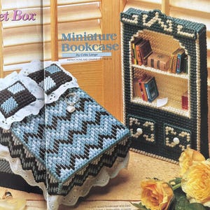 May include: A plastic canvas craft project featuring a miniature bookcase in dark green with cream accents, containing small books on shelves and two drawers. Beside it sits a bed with a blue and black chevron patterned quilt and white lace.