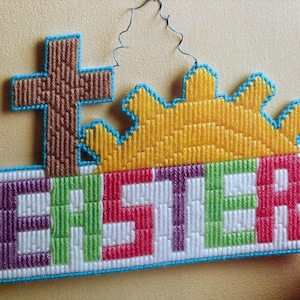 May include: A colorful cross stitch wall hanging with the word "Easter" in a rainbow of colors. A yellow sun with a brown cross is above the word.