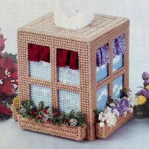 May include: A brown and beige tissue box cover with a window design. The windows have blue and white panes and red and purple curtains. The box has a small planter with green foliage, pine cones, and red berries.